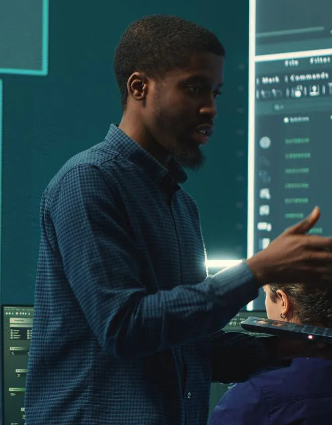 A man in a blue checkered shirt presenting in front of digital screens displaying data and code.