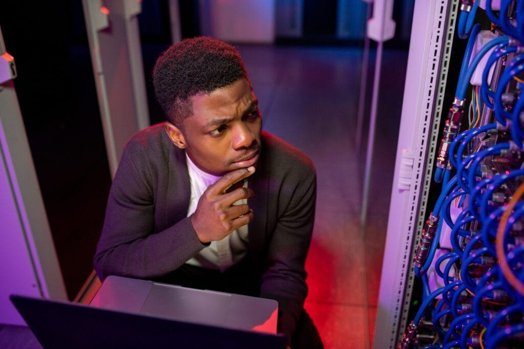 Individual seated with a laptop in a server room, looking thoughtfully at the equipment.