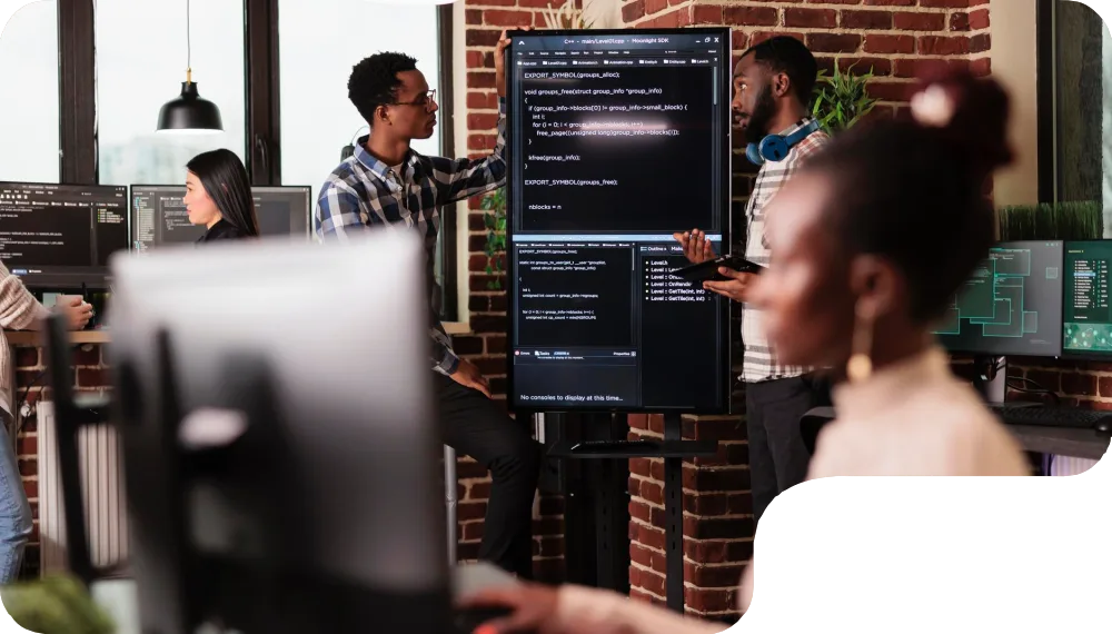 A group of individuals discussing a coding project in a modern workspace with computer screens and a large display.