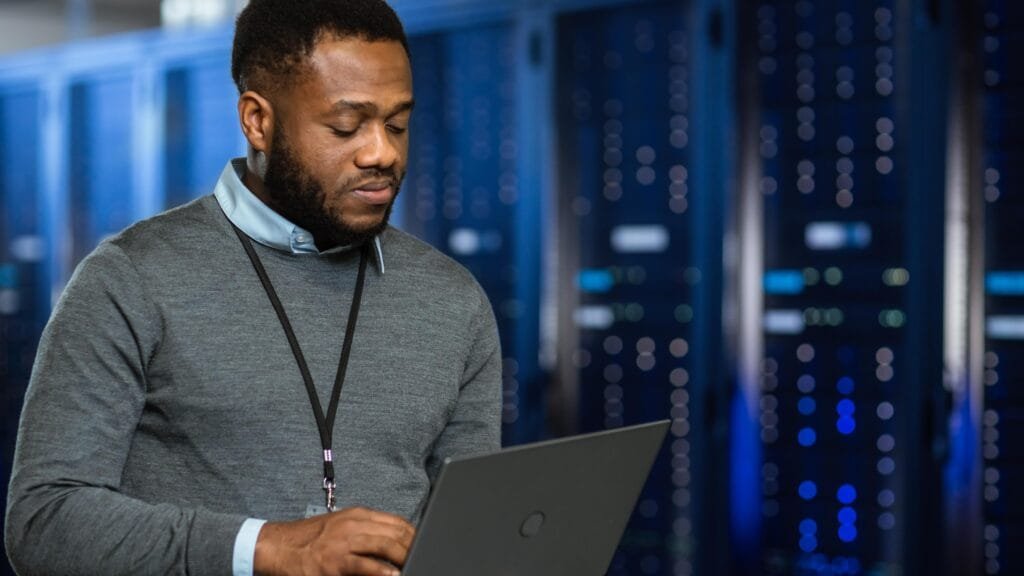Man with a black lanyard around his neck using a laptop in a server room with blue lights.